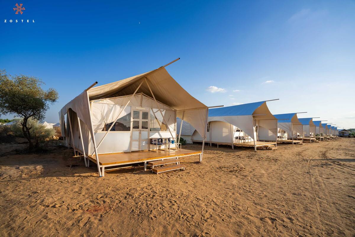 Fileira de tendas no deserto, com um deck de madeira e janela, instalada em solo arenoso com vegetação rasteira.