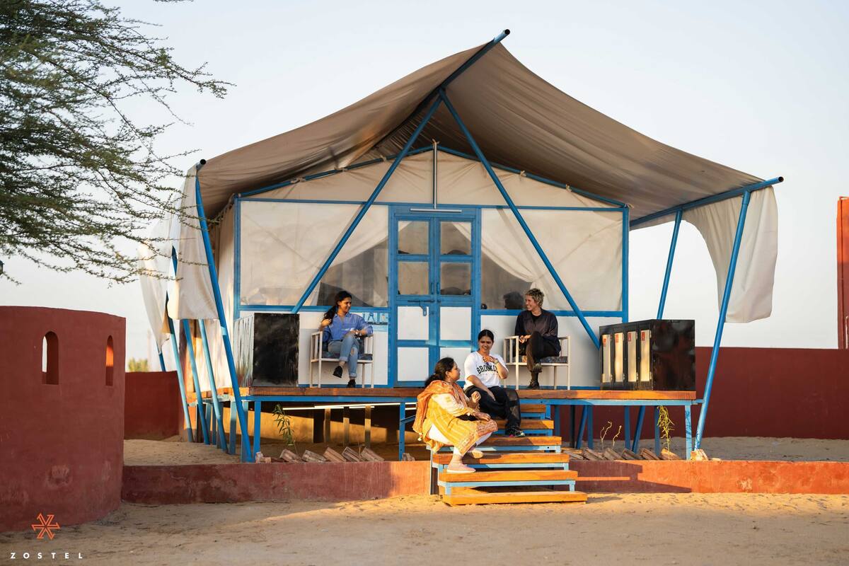 Uma estrutura em forma de tenda, com estrutura azul e toldo bege, apresentando um pequeno alpendre com degraus que levam a ele.