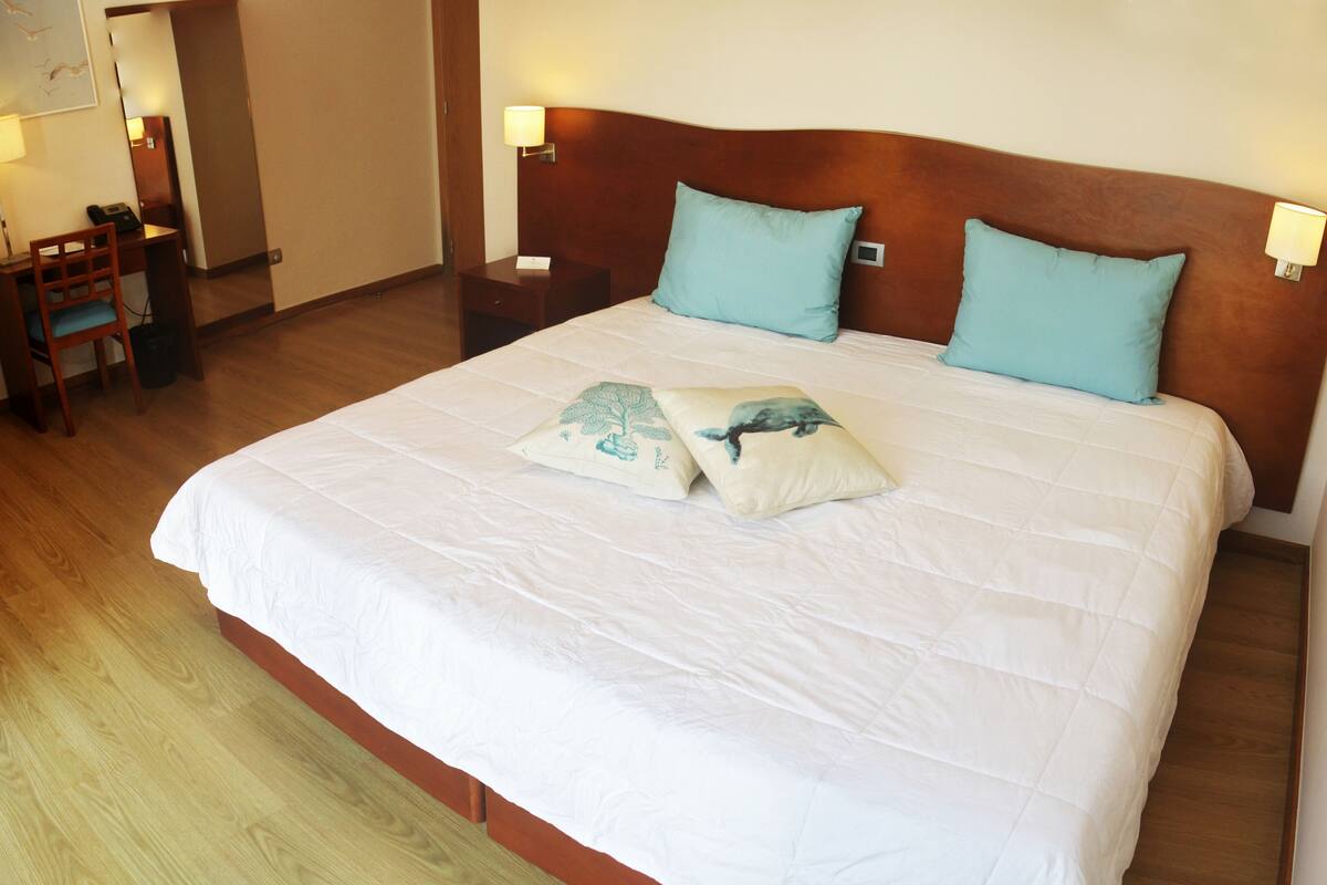 Quarto de hotel com uma cama grande, duas luminárias fixadas na parede, uma mesa de cabeceira de madeira e uma cadeira.
