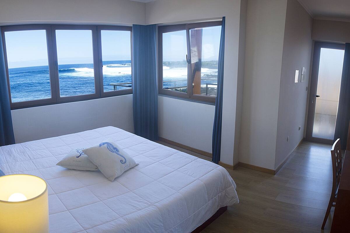 Quarto com cama grande, janela com vista para o oceano e porta que dá para outro espaço.