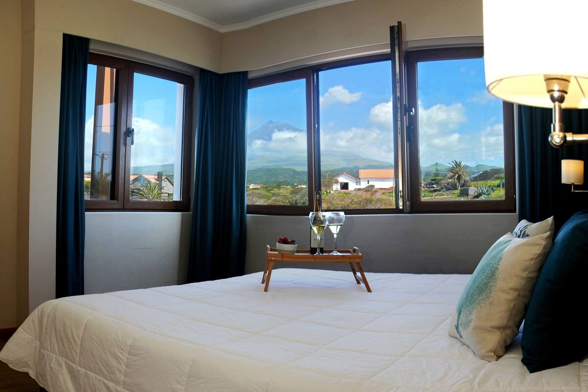 Quarto de hotel com uma cama grande, uma mesa de cabeceira com uma garrafa de vinho e copos, e vista para a paisagem pelas janelas.