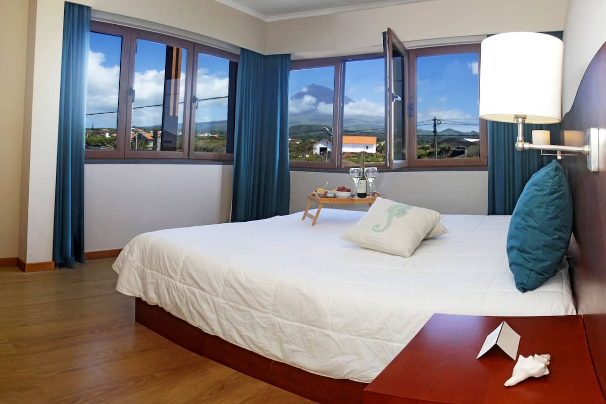 Quarto de hotel com uma cama grande, uma mesinha de cabeceira de madeira, uma mesa de apoio com abajur e vista para a paisagem pelas janelas.