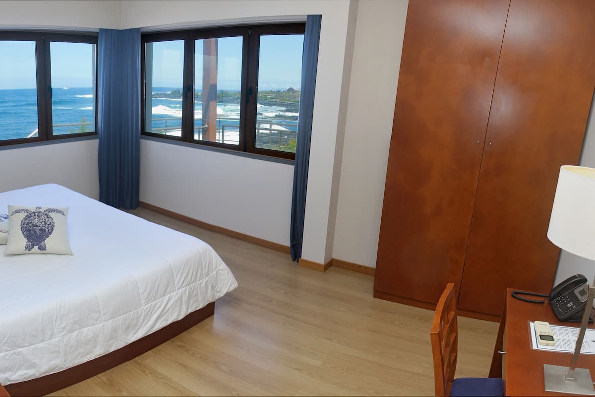 Quarto de hotel com cama, escrivaninha com telefone e grandes janelas com vista para o oceano.