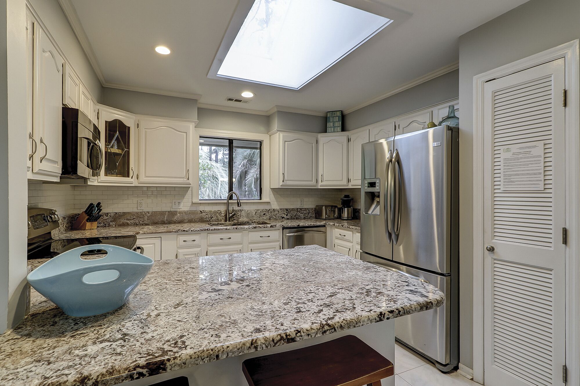 A kitchen with white cabinets, a stainless steel refrigerator, a granite countertop, and a skylight.