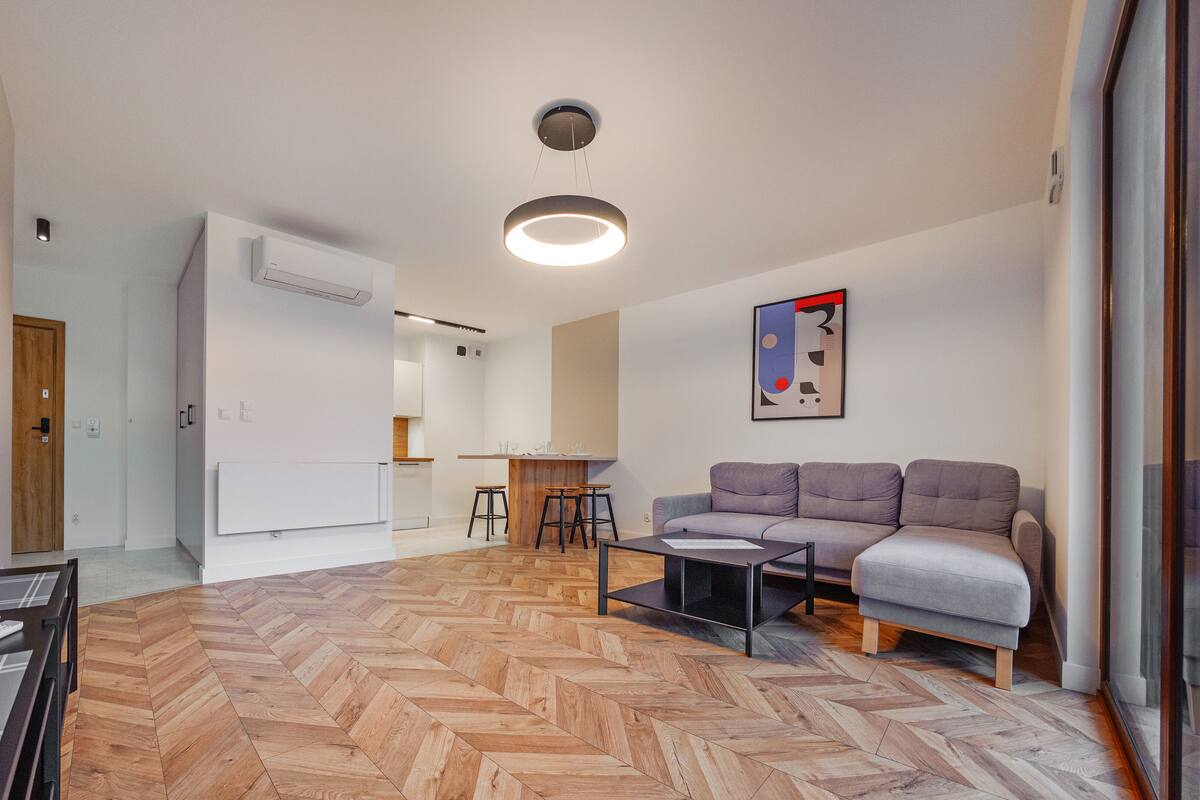 A modern living room with a herringbone wood floor, a grey sofa, a coffee table, and a flat-screen TV mounted on the wall.