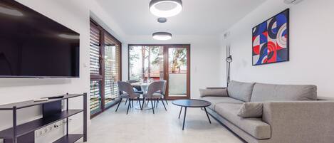 A modern living room with a flat-screen TV, a grey sofa, a dining table with chairs, and a large window with blinds.