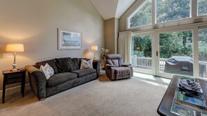 A living room with a dark brown sofa, a grey armchair, and a glass coffee table with a magazine. There are two lamps on side tables, a framed picture on the wall, and a large window with a view of greenery. - 17 Cartgate - Palmetto Dunes - 4 Bed 4 Bath Private Pool -Hilton Head Rentals (Hilton Head Island)