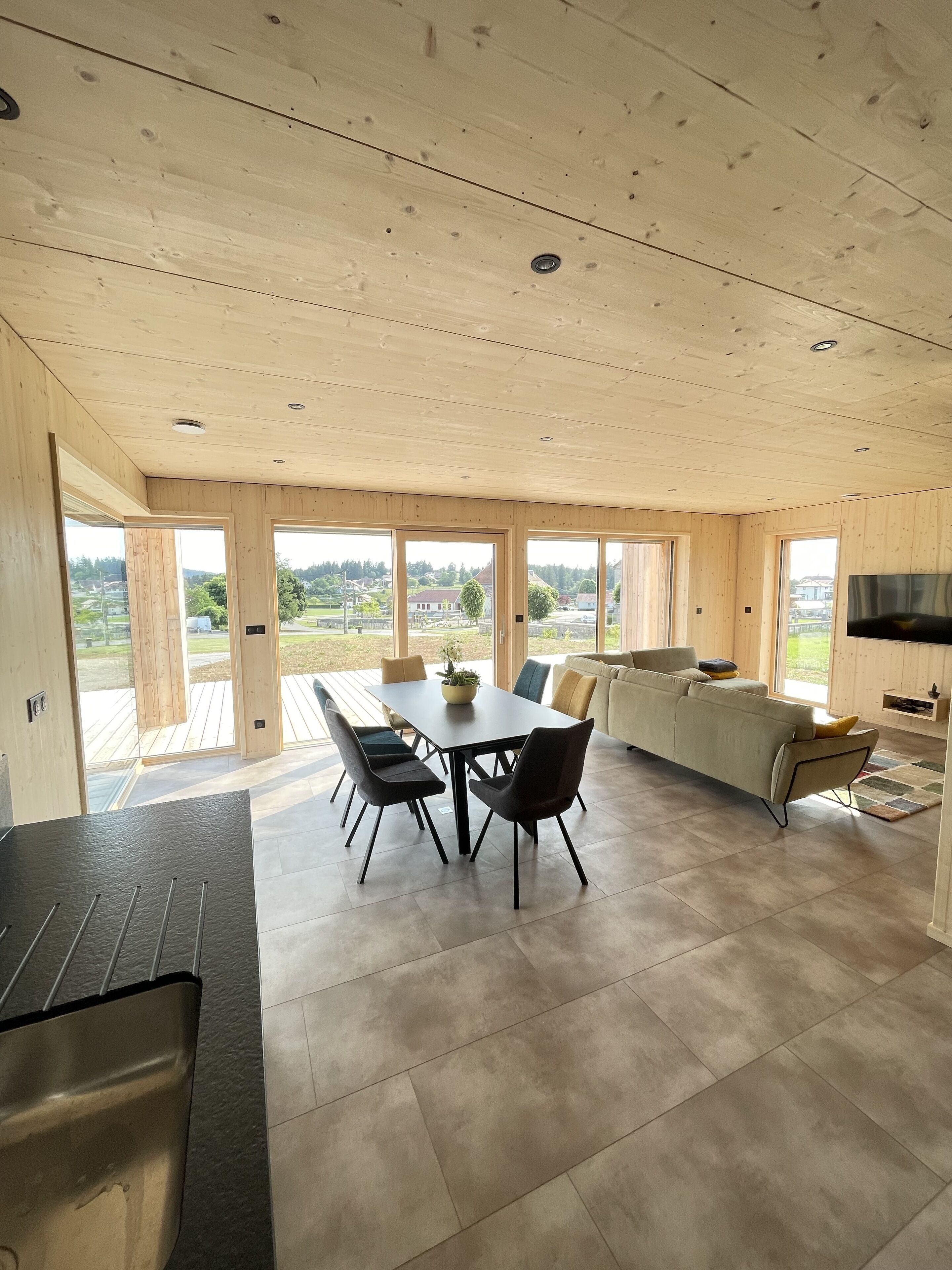 A spacious room with a wooden ceiling, large windows, a dining table with chairs, a sofa, and a TV.