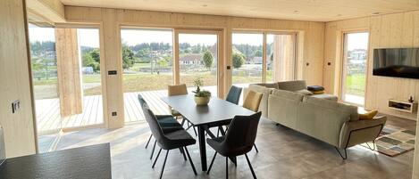A spacious room with a wooden ceiling, large windows, a dining table with chairs, a sofa, and a TV.