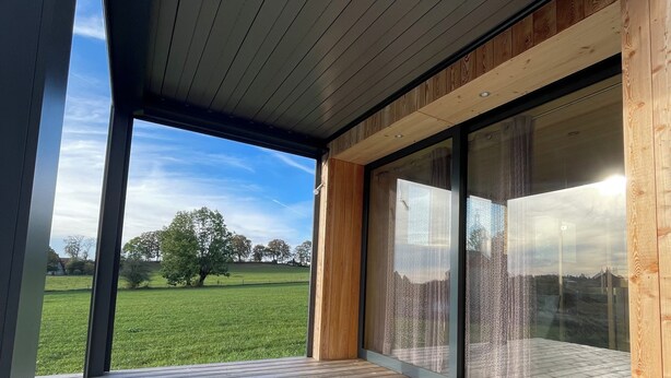 Un bâtiment moderne doté d’une terrasse en bois et de grandes portes vitrées donnant sur une zone verdoyante.