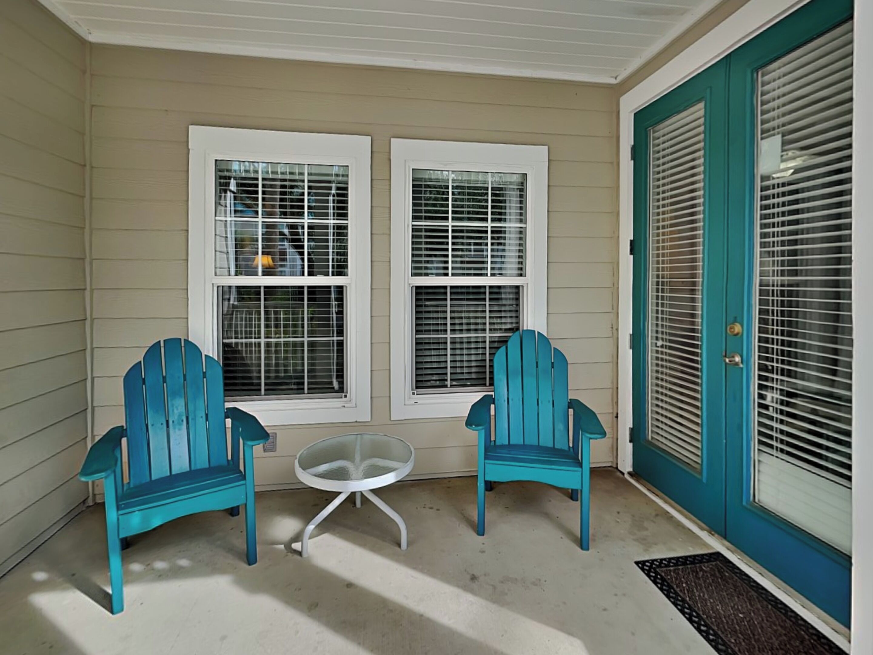 A porch with two blue Adirondack chairs, a small glass table, and a door with teal shutters.