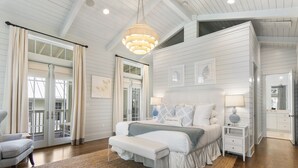 A spacious bedroom with a large bed, a bench, a nightstand, and a dresser. There is a large window with curtains, a chandelier, and artwork on the walls. - Home Across from Beach Access w/ Pool (Santa Rosa Beach)