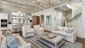 A spacious living room with white furniture, a glass coffee table, and a chandelier. There is a kitchen area with white cabinets and a dining table. - Home Across from Beach Access w/ Pool (Santa Rosa Beach)