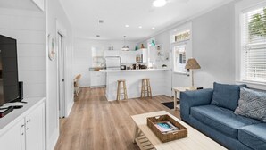 A modern kitchen with white cabinets, a central island with stools, a dining area with a blue sofa, and a large window with blinds. - Renovated 30A Bungalow 5 Min Walk to Beach!! (Santa Rosa Beach)