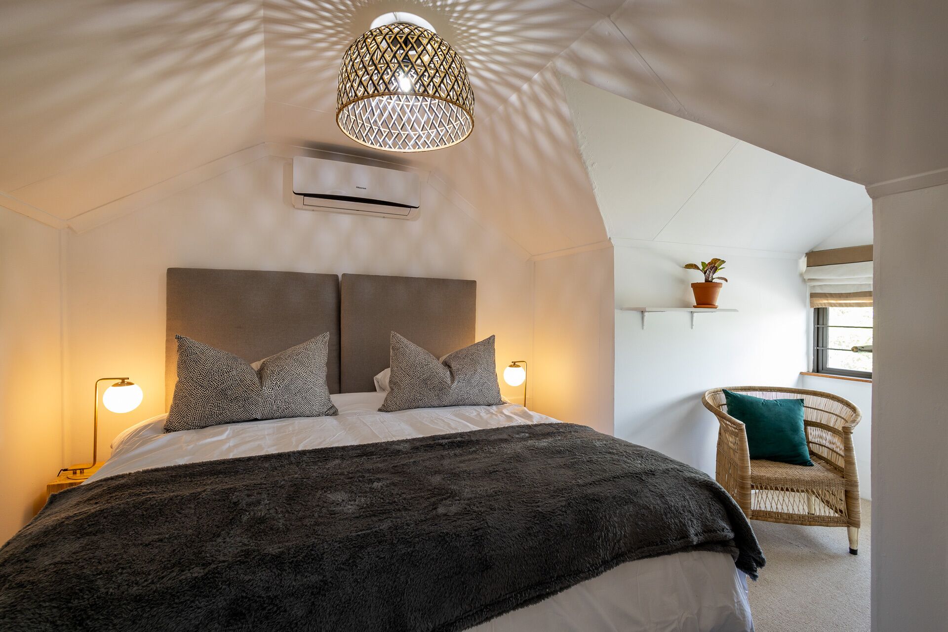 A bedroom with a large bed, two bedside lamps, a hanging light fixture, a potted plant, and a wicker chair.