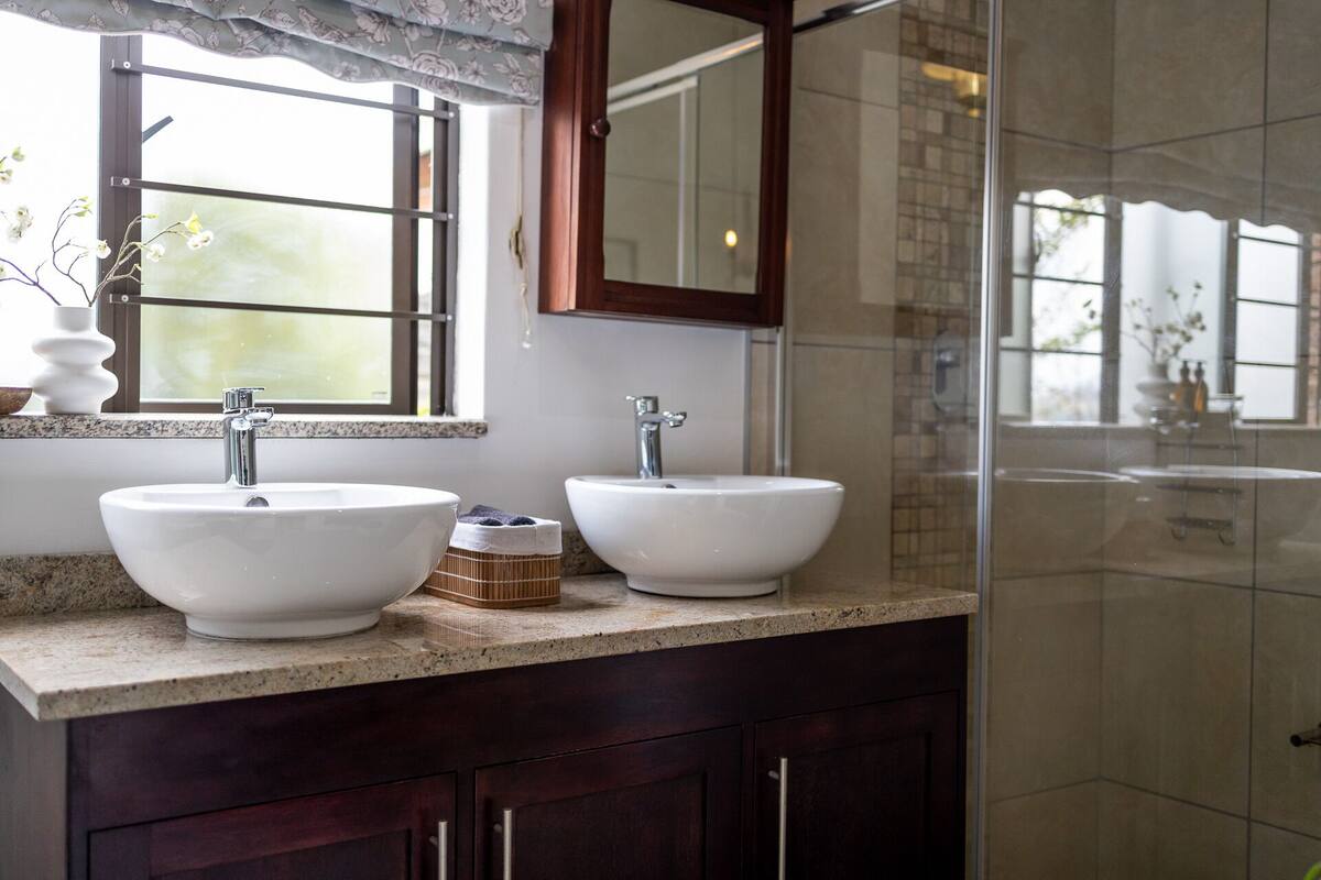 Une salle de bain avec deux lavabos blancs, un miroir au-dessus, et une fenêtre ornée d’un rideau à motifs floraux.
