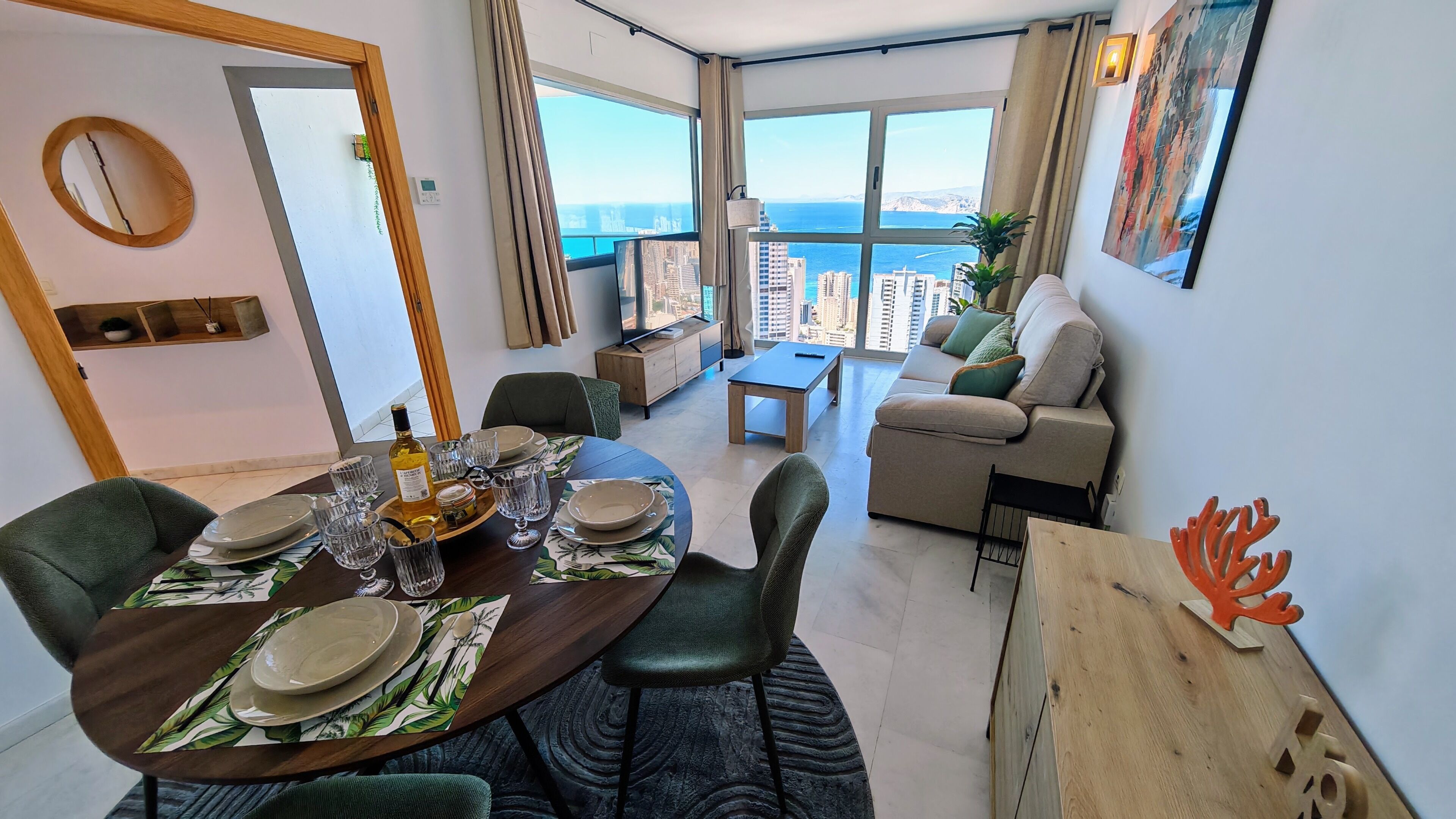 Un soggiorno moderno con zona pranzo, ampie finestre vista città e mare, un divano e un tavolo da pranzo per quattro persone.