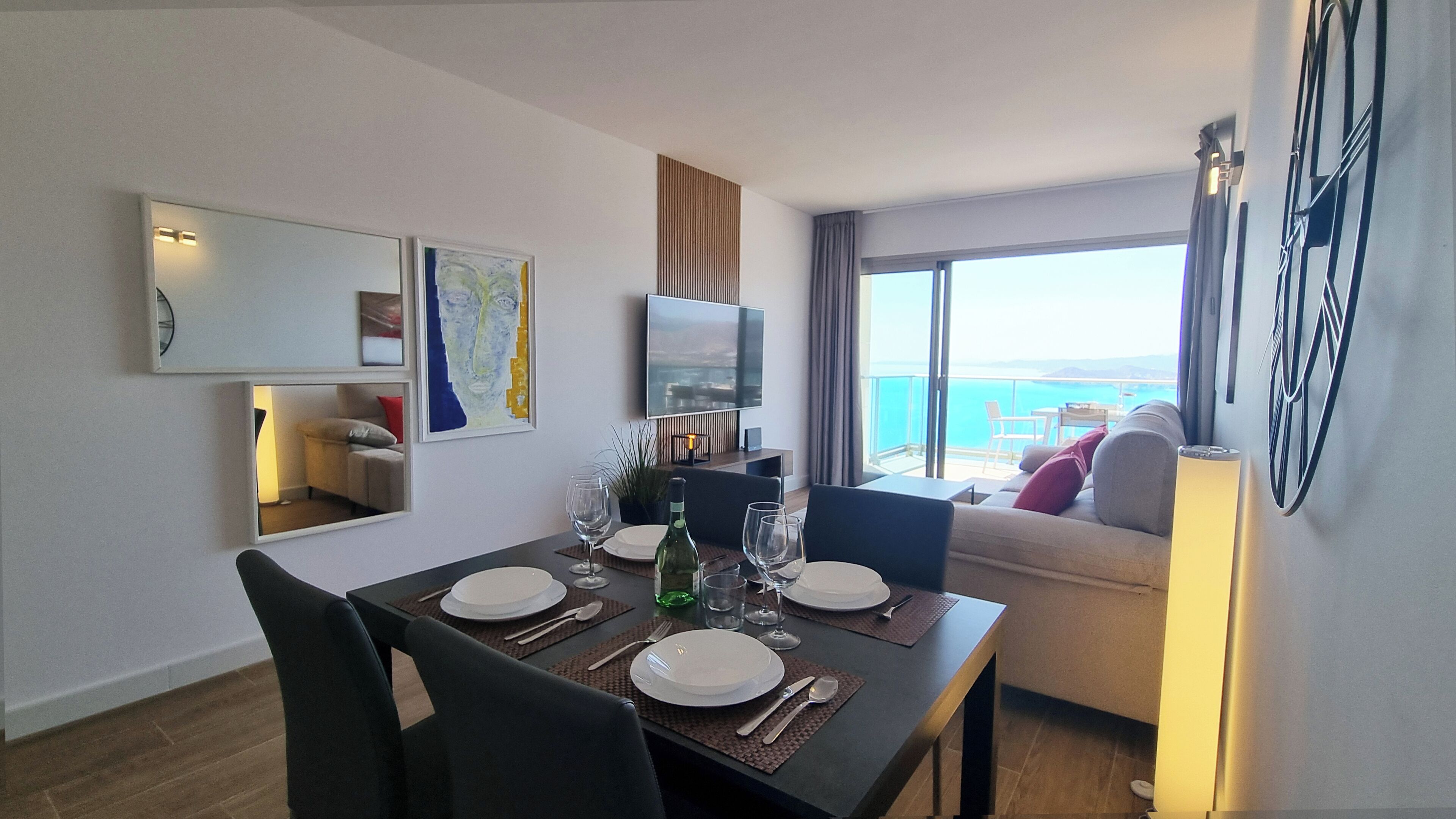 A dining area with a table set for four, a sofa, a TV, and a view of the sea.