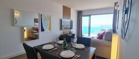 A dining area with a table set for four, a sofa, a TV, and a view of the sea.