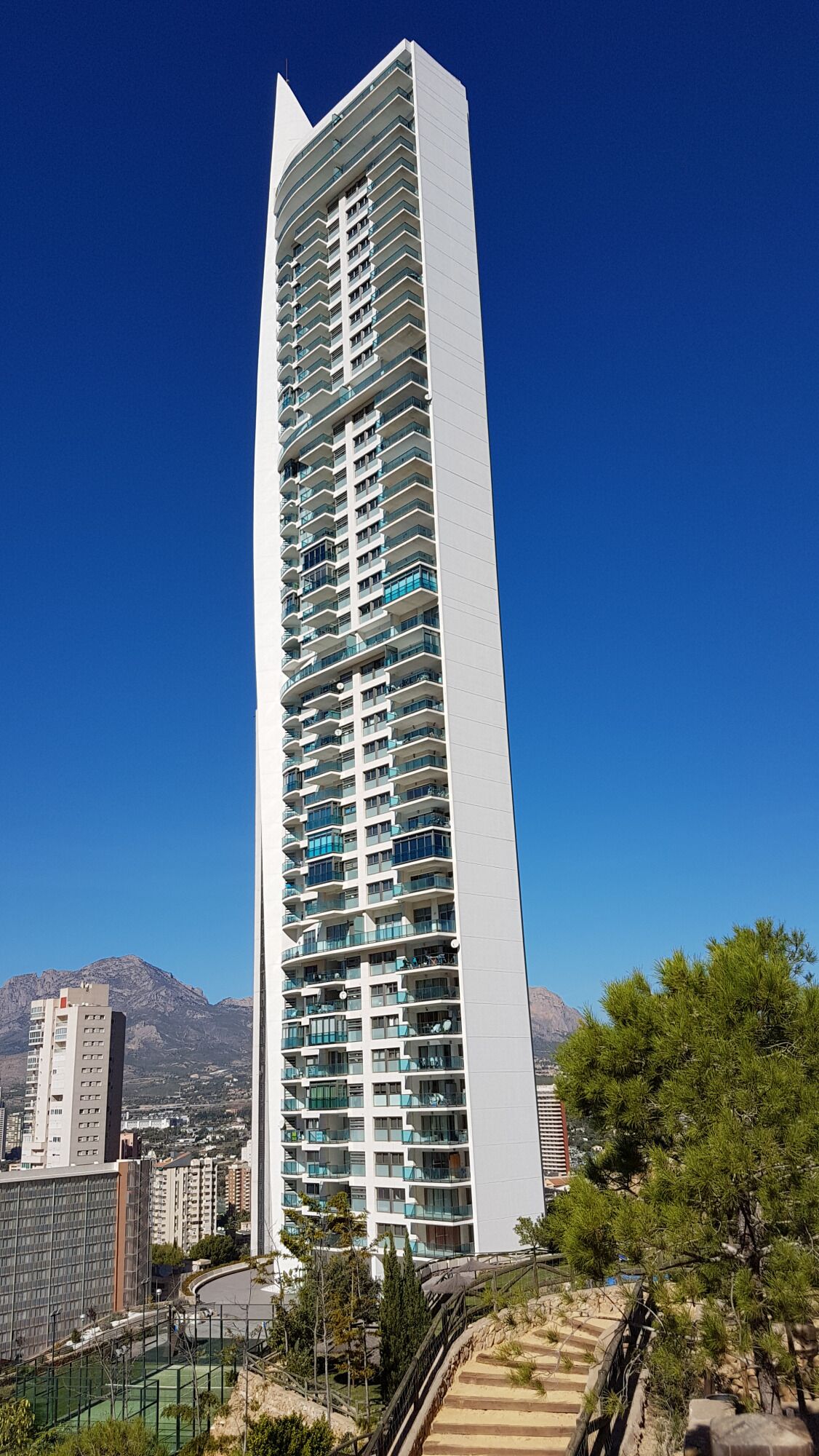 A tall, modern building with multiple balconies, surrounded by greenery and mountains in the background.