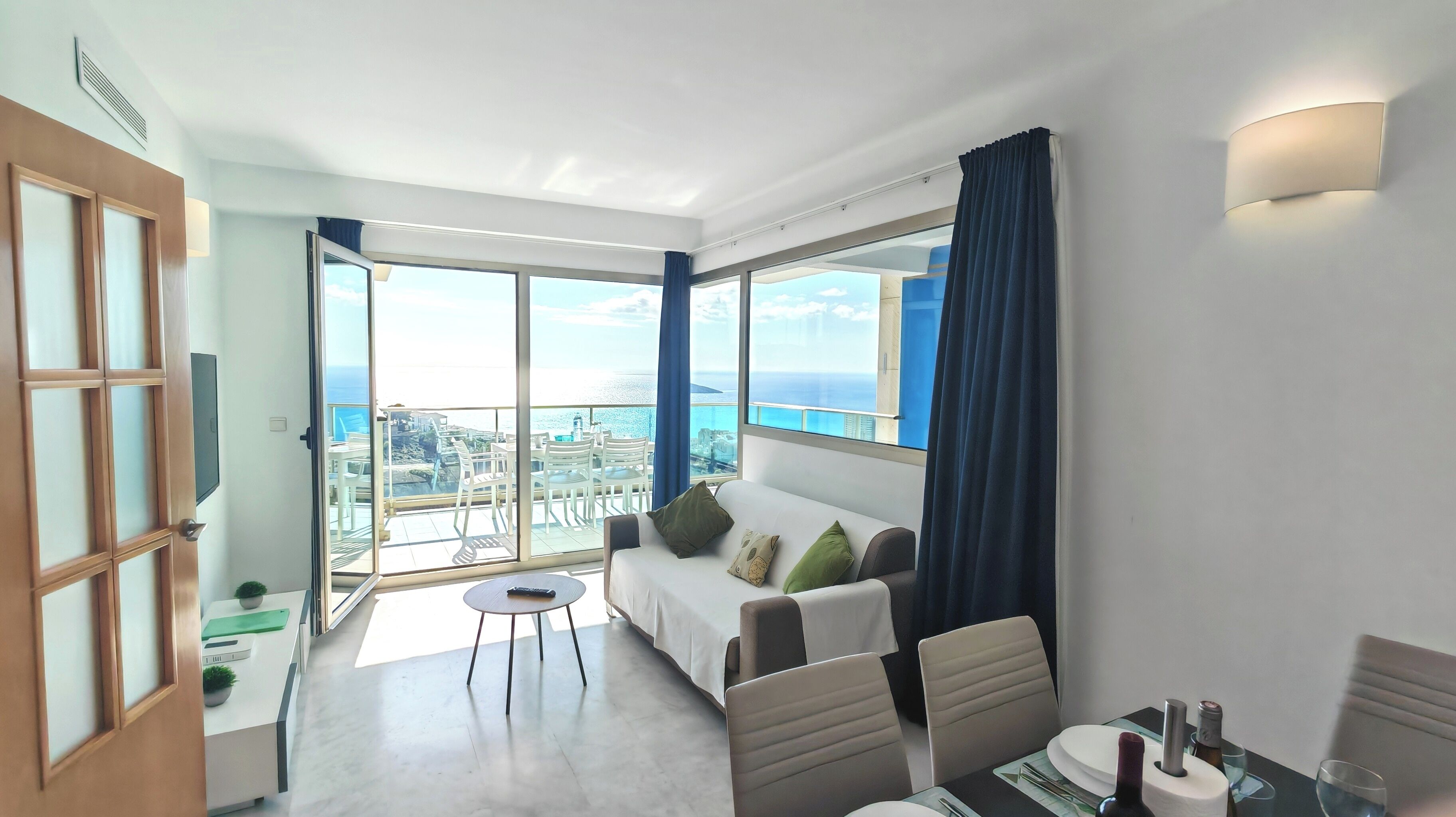 Una habitación de hotel moderna con balcón, sofá, mesa de comedor y televisión.