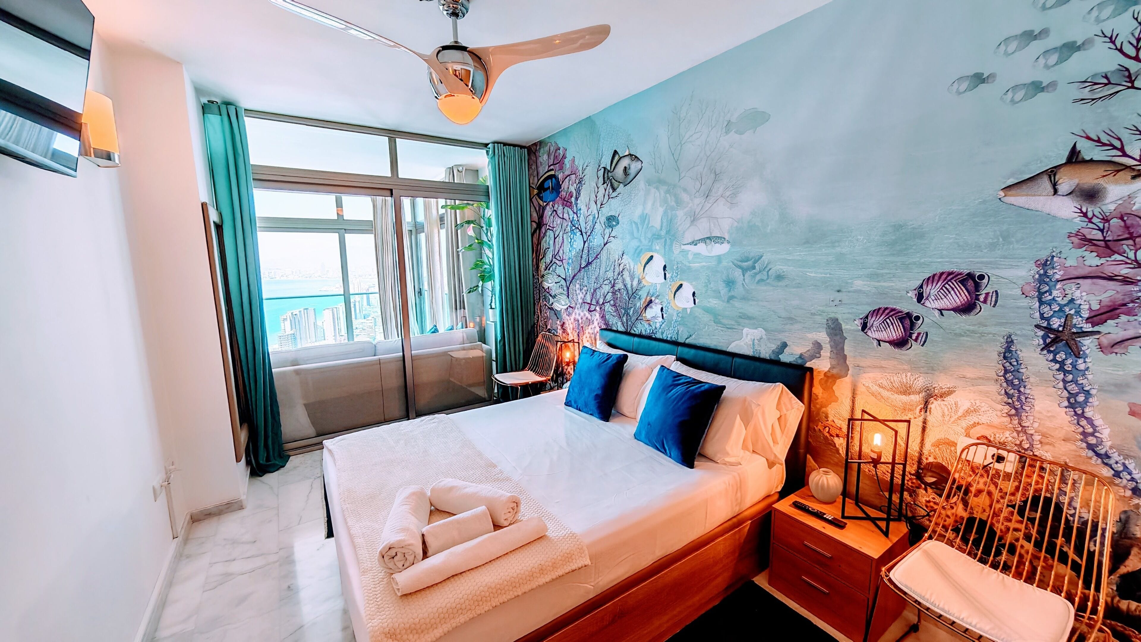 A hotel room with a bed, a ceiling fan, a nightstand, a wall mural, and a view of the city.