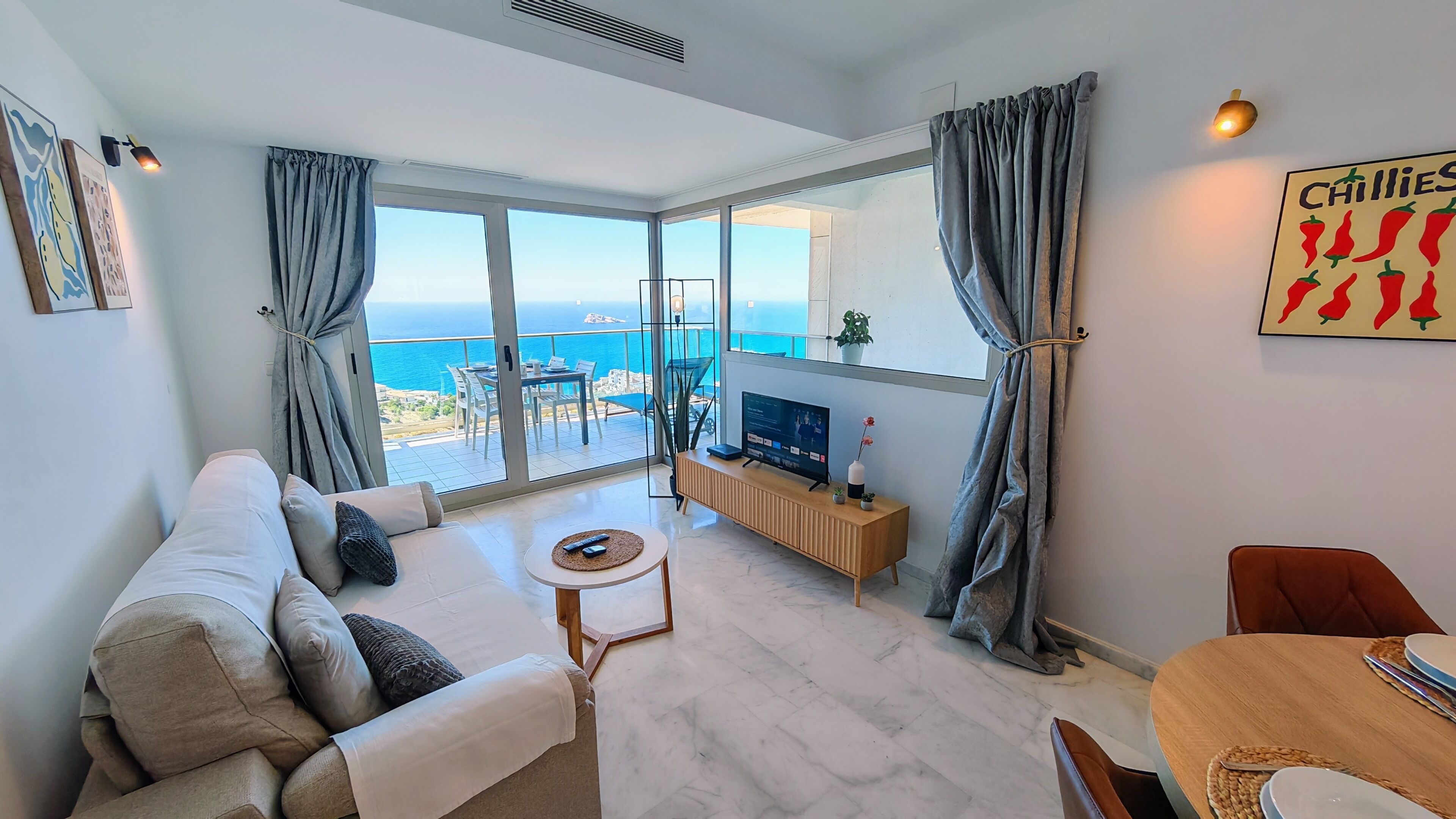 A modern living room with a sofa, a coffee table, and a TV. There is a balcony with a view of the sea.