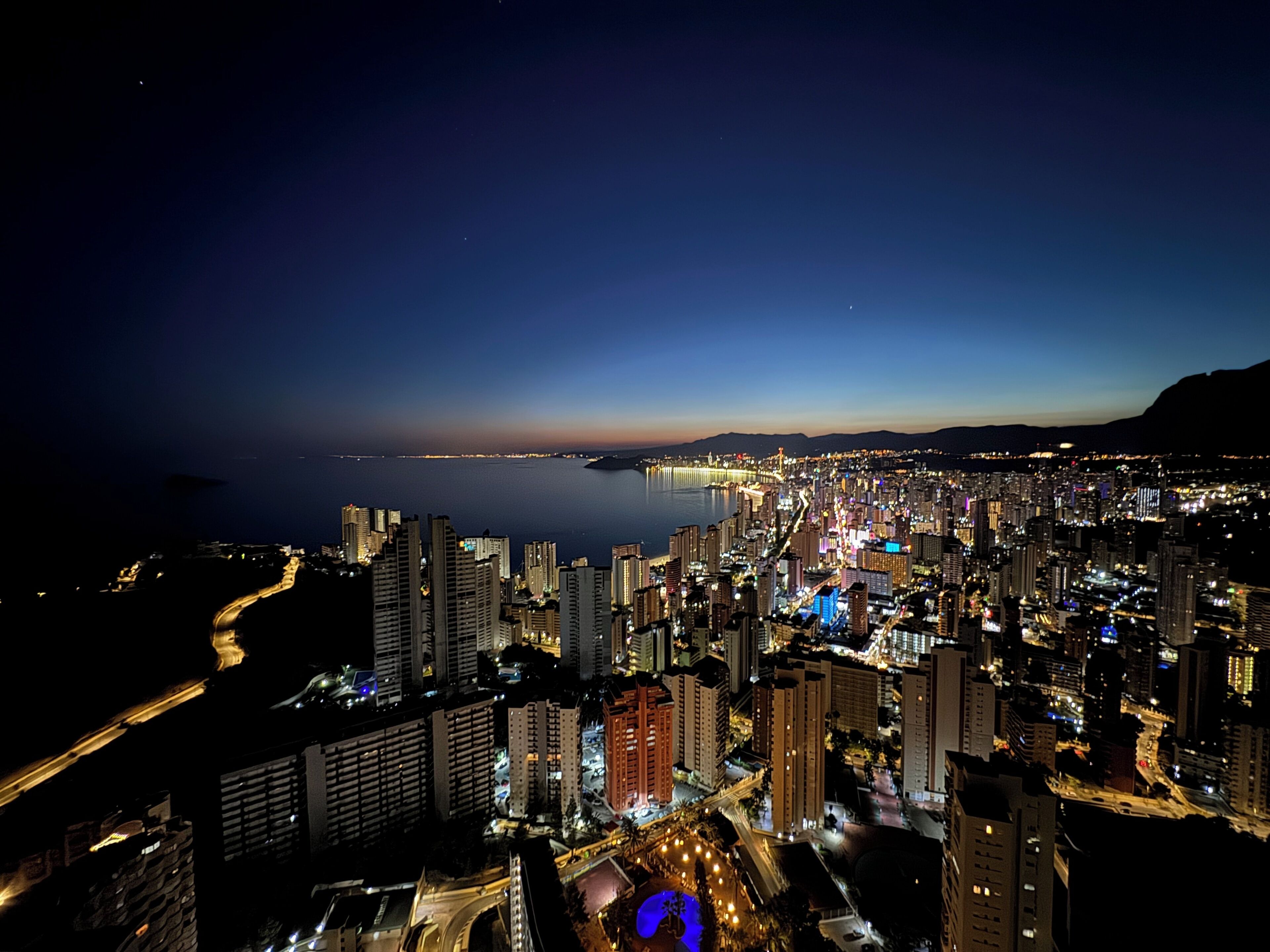 A nighttime cityscape with high-rise buildings, a waterfront, and a bridge.