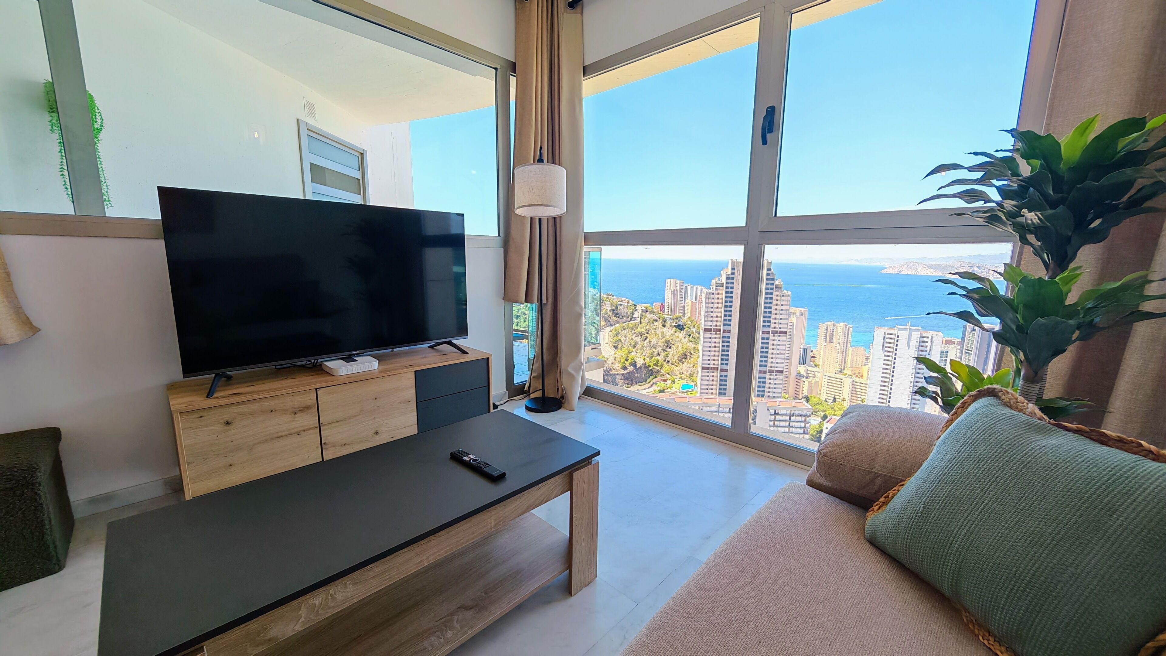 A modern living room with a large TV, a wooden coffee table, a sofa, and a view of the city and sea.