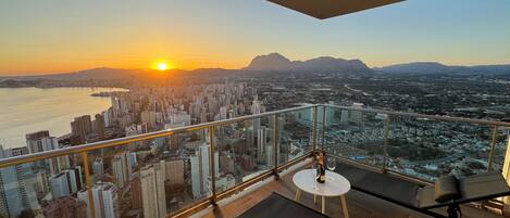 A balcony with a city view at sunset, a table with a bottle and glass, and a chair.