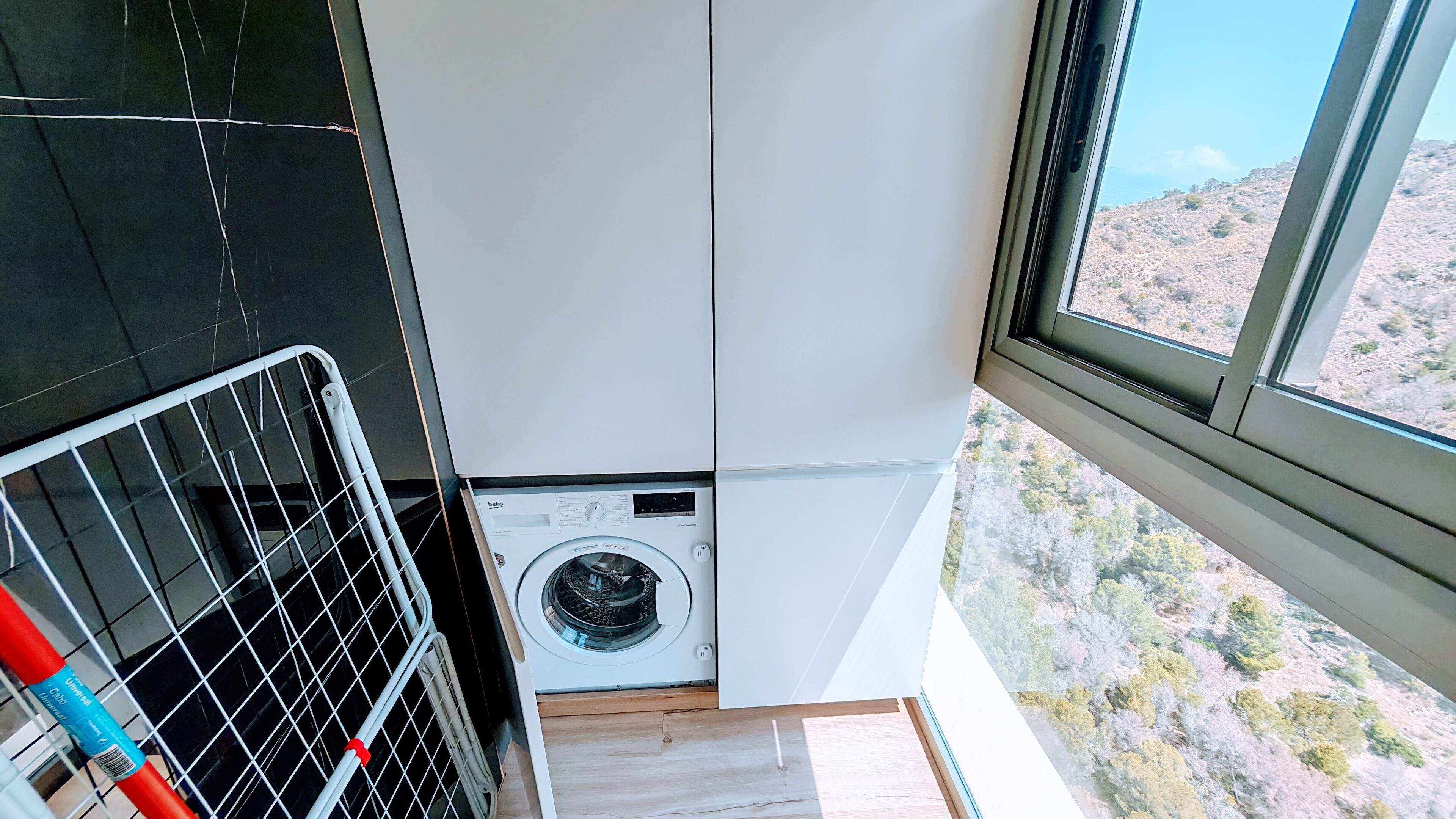 Een kleine kamer met een wasmachine, een metalen rek en een raam met uitzicht op een glooiend landschap.