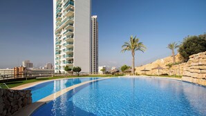 A swimming pool with a curved edge, a tall building in the background, and a rocky landscape with palm trees. - Benidorm sky - High-rise apartments (Benidorm)