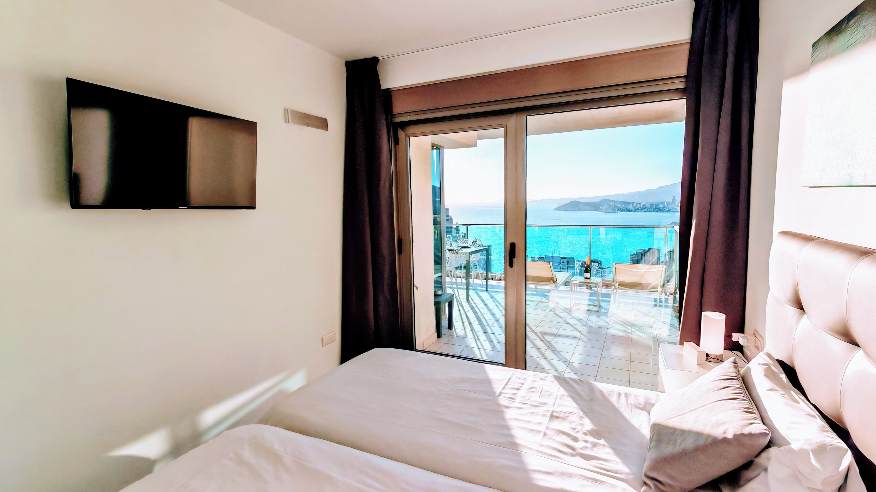 A hotel room with a bed, a TV, and a balcony with a view of the sea.