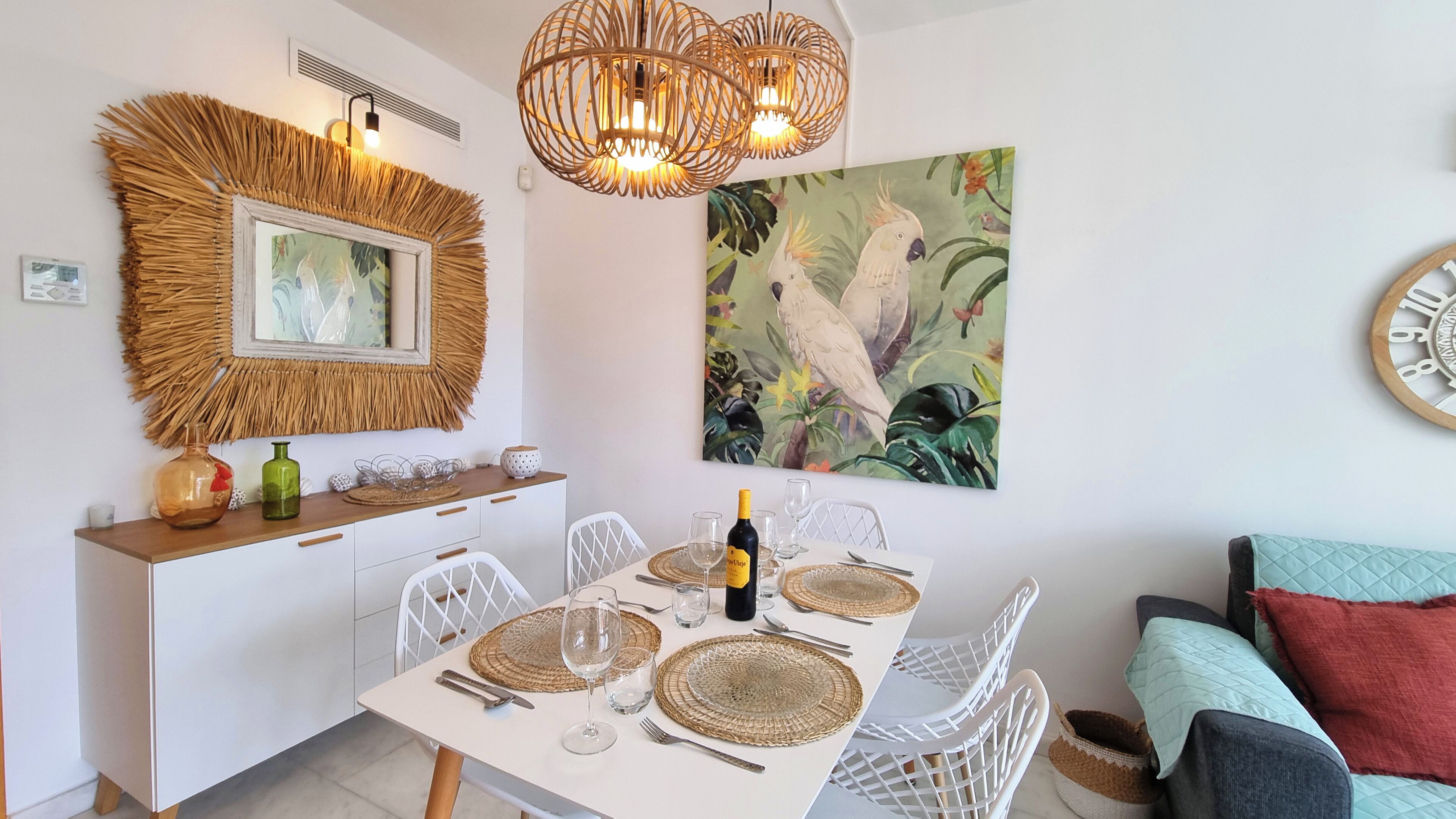 Een eetgedeelte met een witte tafel voor zes personen, omringd door witte stoelen. Op de tafel staan een fles wijn en glazen. Daarboven hangt een ingelijste foto van een vogel.