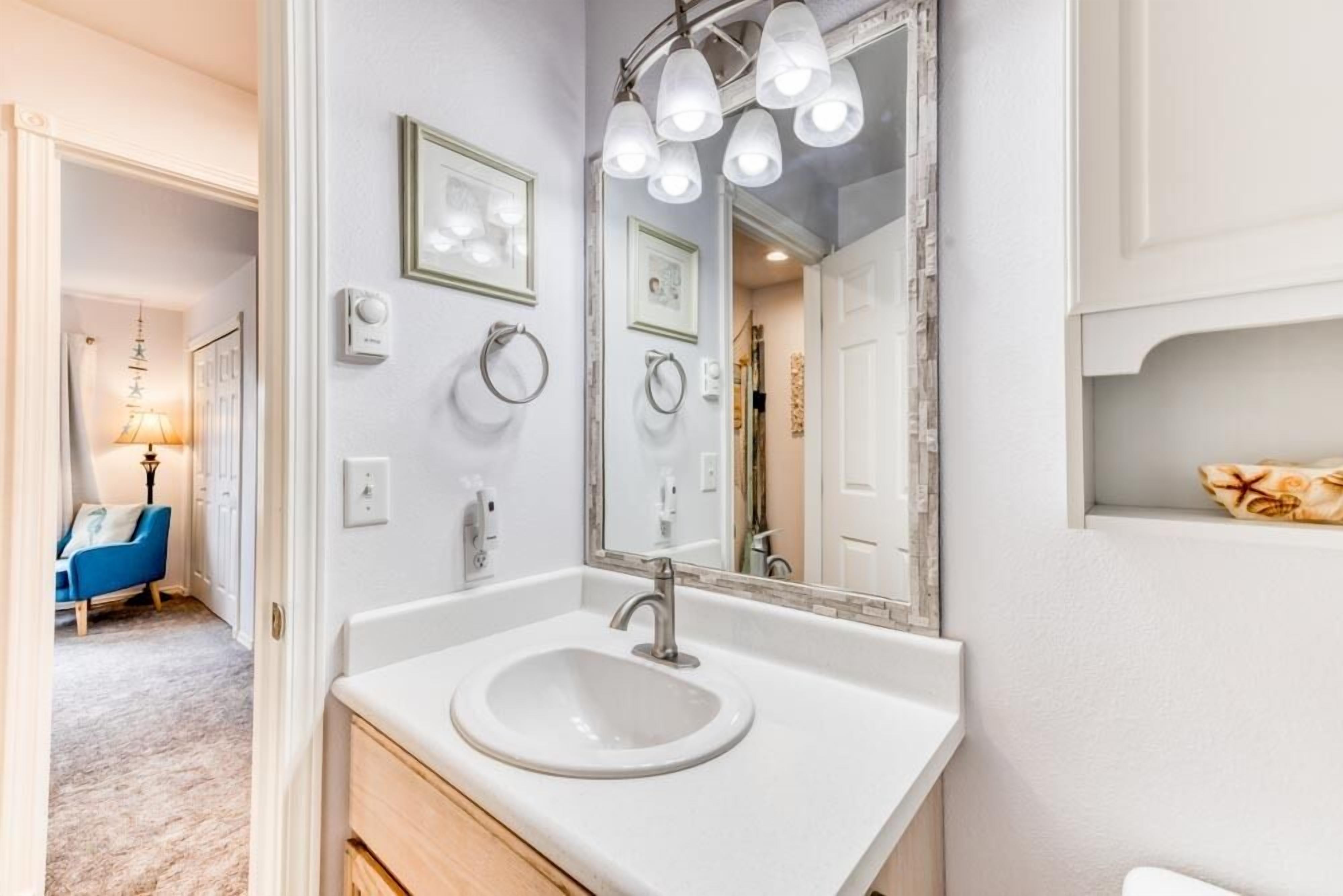 Une salle de bain avec un lavabo blanc, un grand miroir avec éclairage intégré et une chaise bleue à l’arrière-plan.