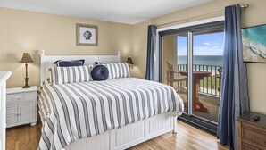 A bedroom with a bed, a nightstand, a lamp, a framed picture, and a balcony with a view of the ocean.