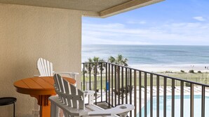 A balcony with a table and chairs, overlooking a beach and a pool.