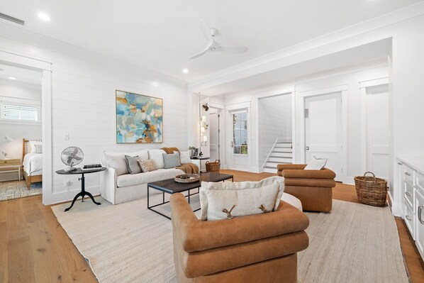 A modern living room with a white sofa, a brown armchair, a coffee table, and a fan. There is a painting on the wall and a staircase leading to another room.