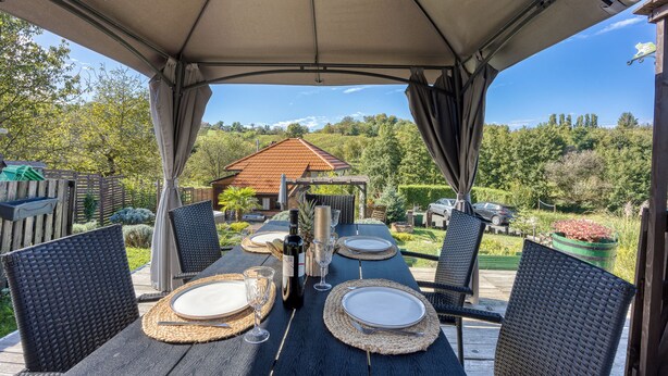 An outdoor dining area with a covered table set for four, surrounded by wicker chairs, overlooking a garden and a house with a red roof. - Romantic Haven With Hot Tub (Beretinec)