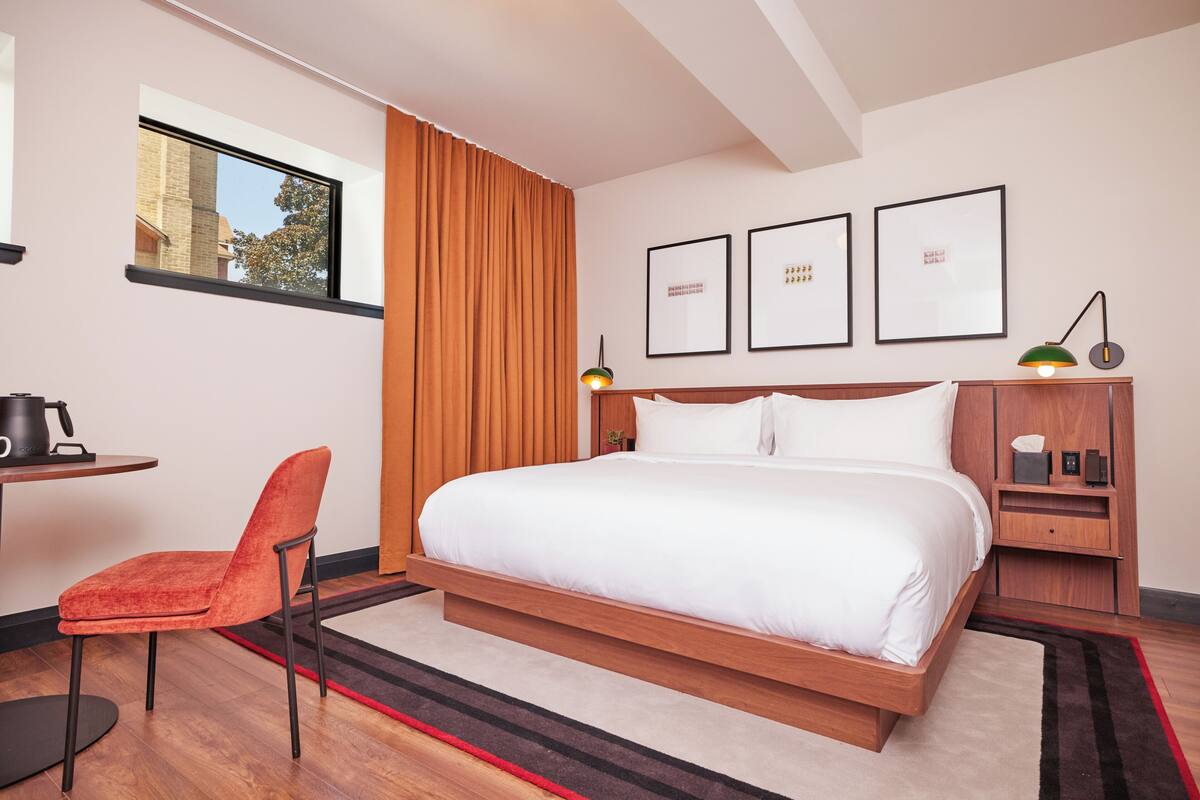 A modern hotel room with a large bed, a desk, a chair, and a nightstand with a lamp.