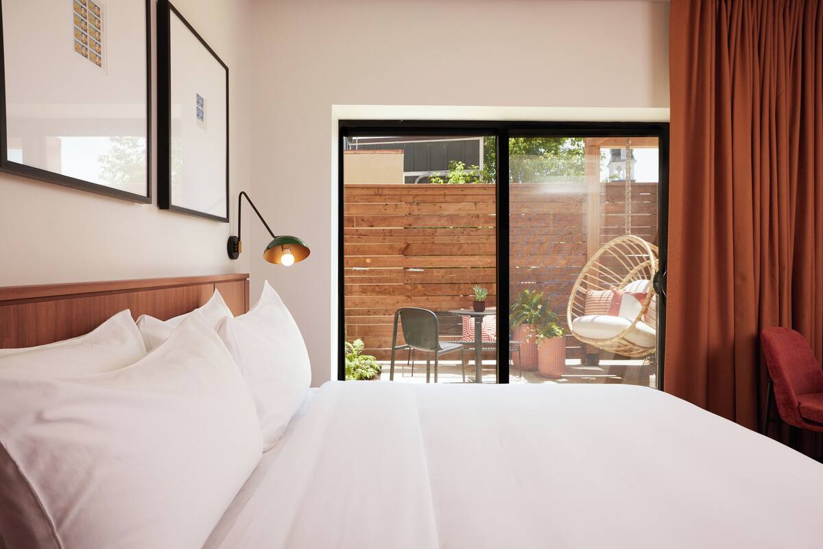 A hotel room with a large bed, a wooden headboard, a framed picture on the wall, a table with chairs, and a view of the outside through a sliding door.