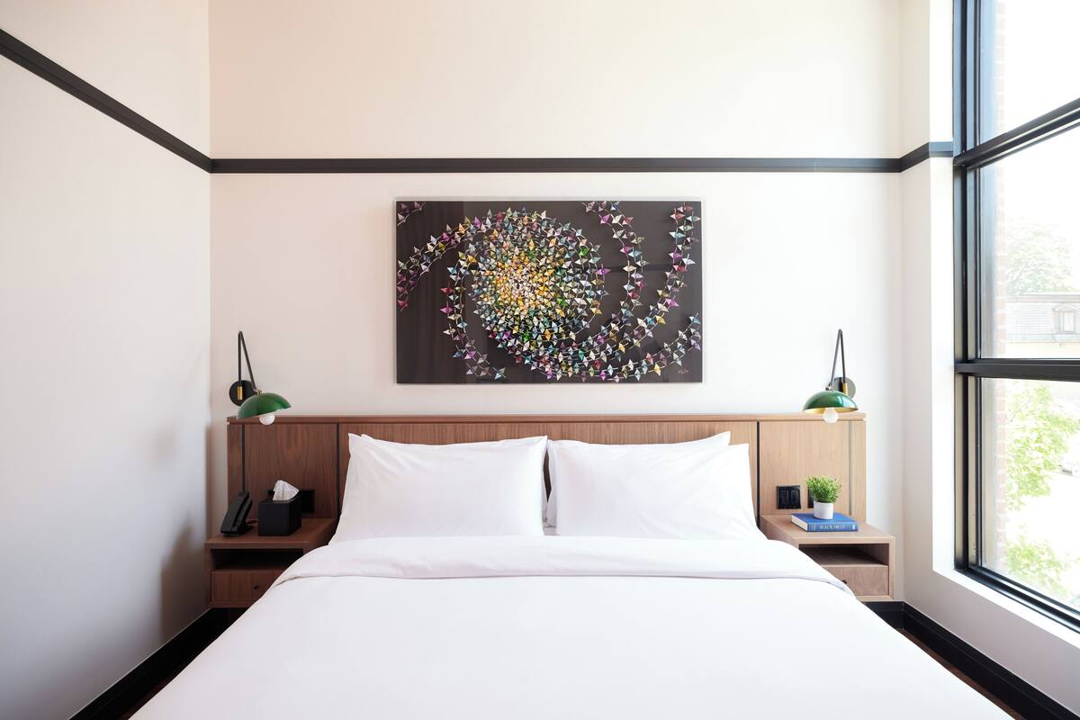 A hotel room with a bed, a nightstand with a lamp, a small table with a plant, and a wall art piece.