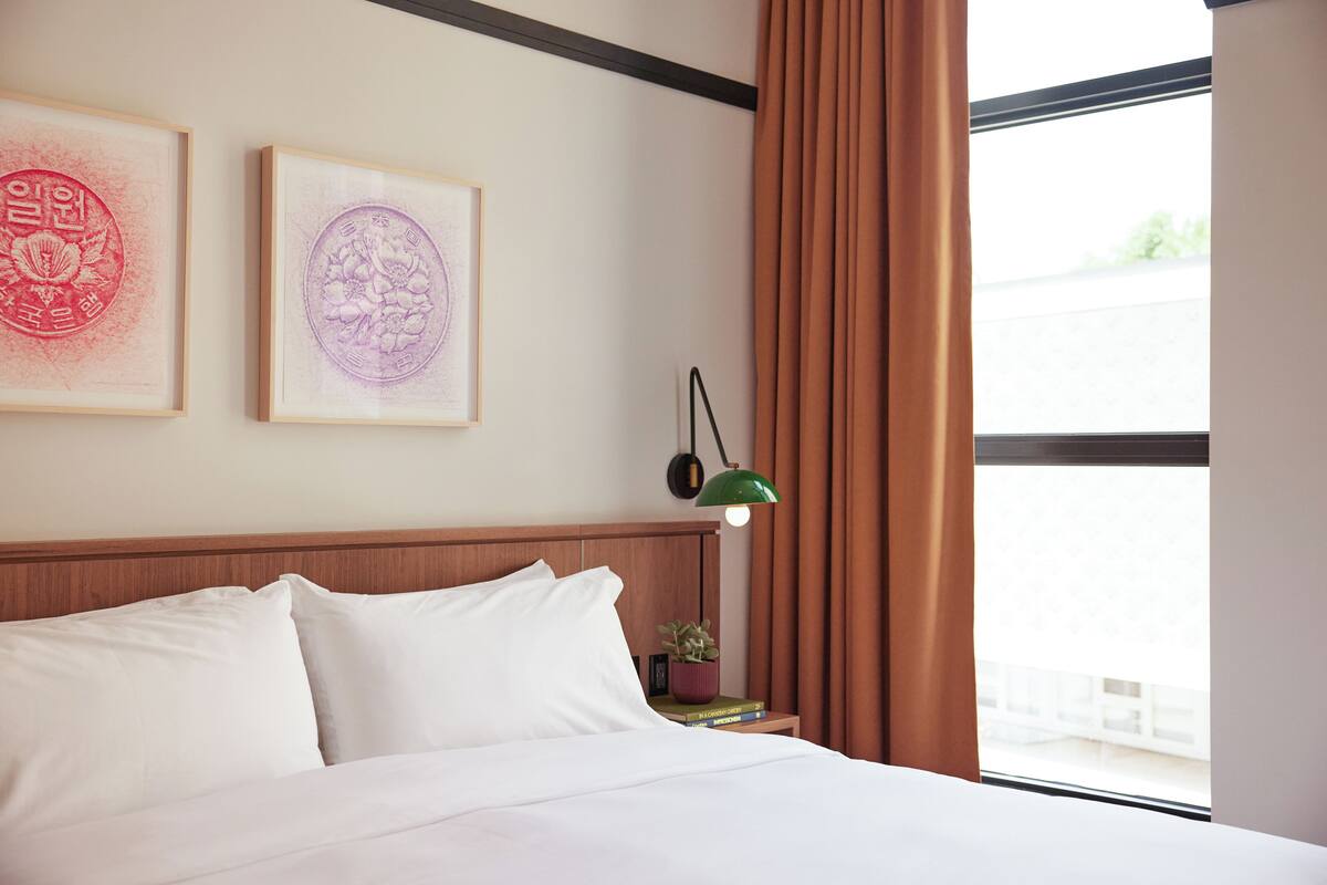 A neatly made bed with white linens, a green wall-mounted lamp, and two framed artworks above it.