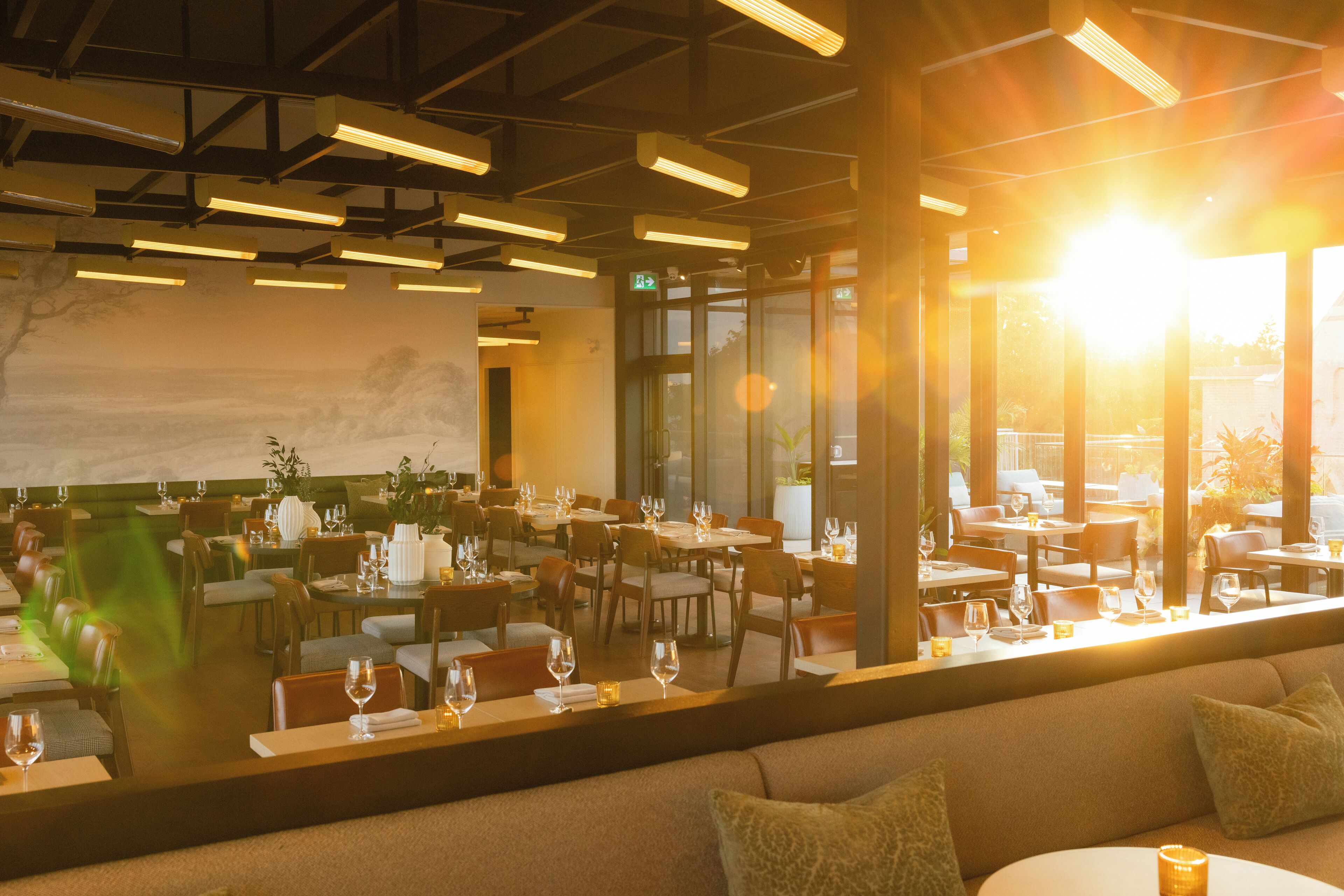 Un restaurant doté de nombreuses tables et chaises, d’une grande fenêtre offrant une vue sur l’extérieur, et d’un plafond moderne avec des poutres apparentes et des luminaires.