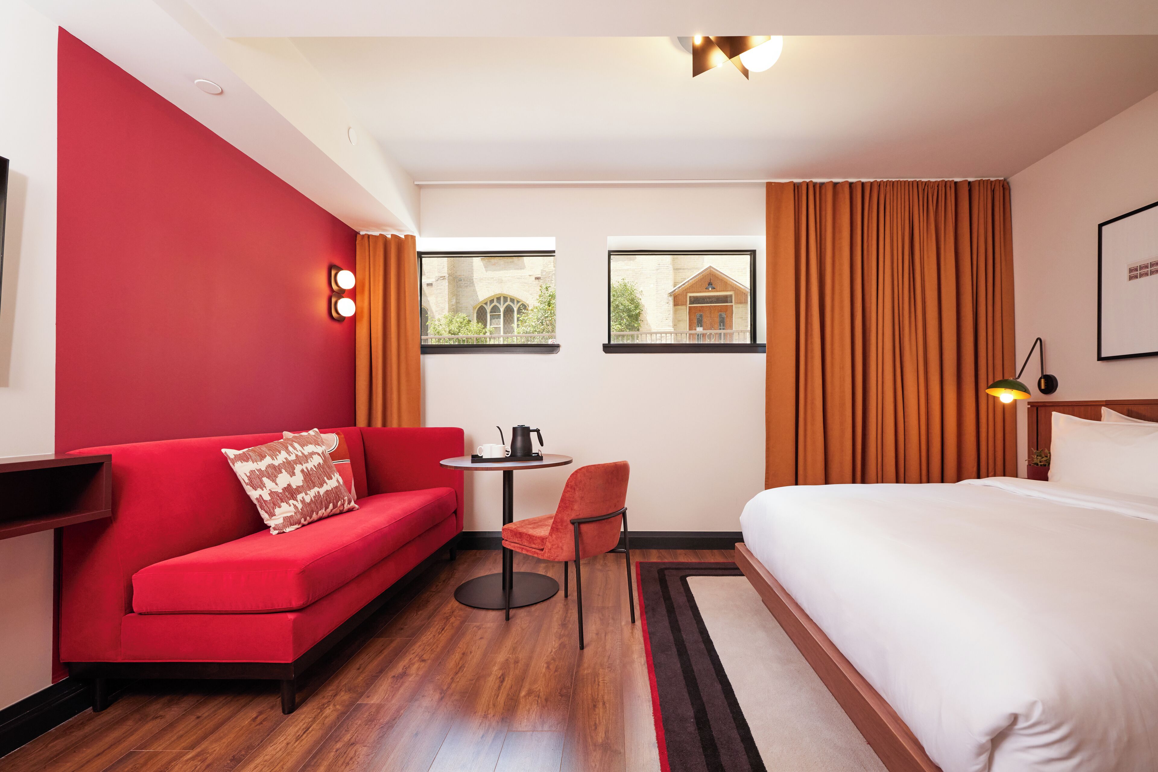 Une chambre d’hôtel avec un canapé rouge, une petite table ronde avec une bouilloire, une chaise, un lit et deux fenêtres avec des rideaux.