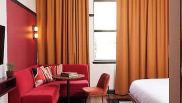 A modern hotel room with a red sofa, a small table, a single bed, and large windows with curtains.