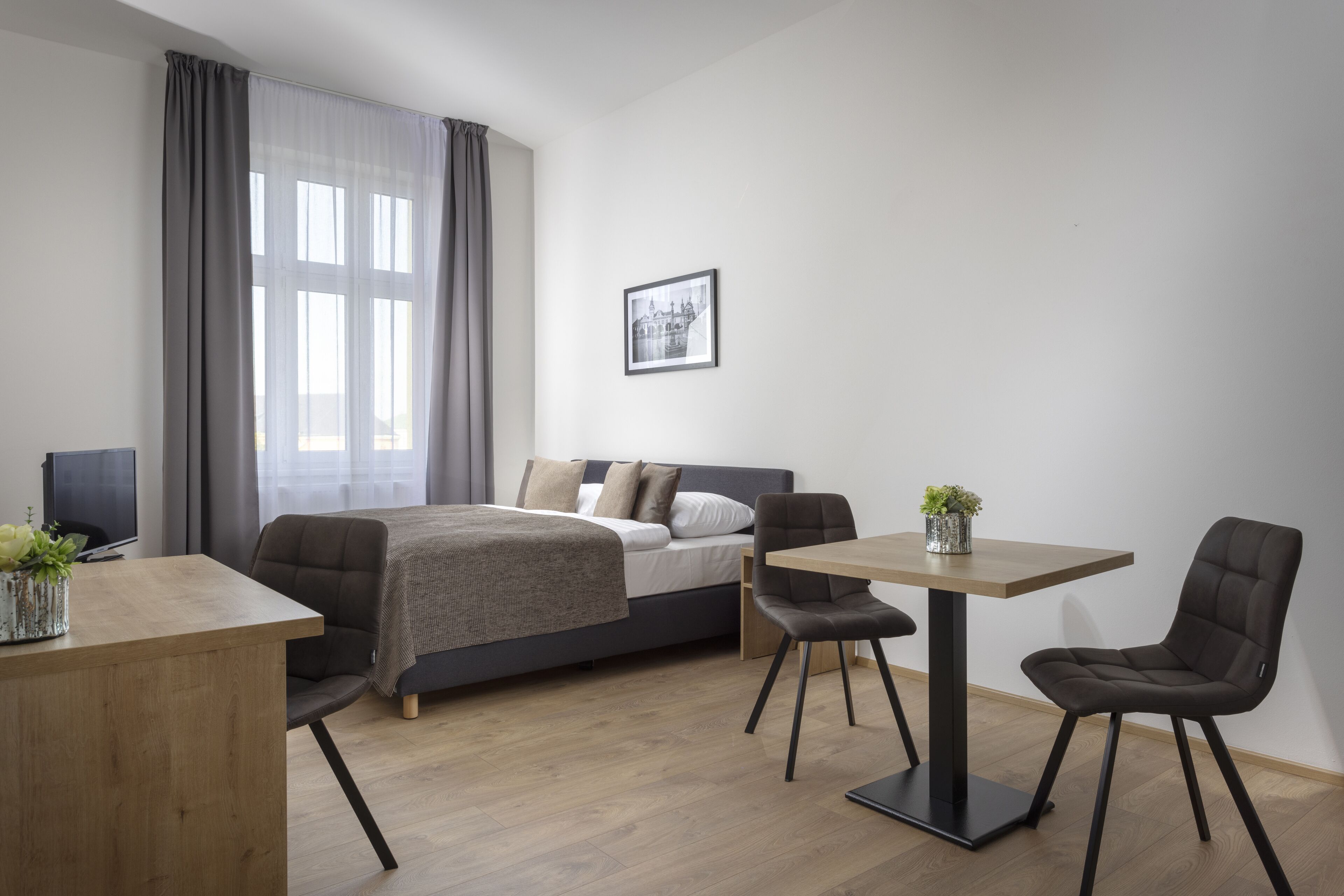 Moderni hotellihuone, jossa on sänky, työpöytä, kaksi tuolia, televisio ja seinällä kehystetty taulu.