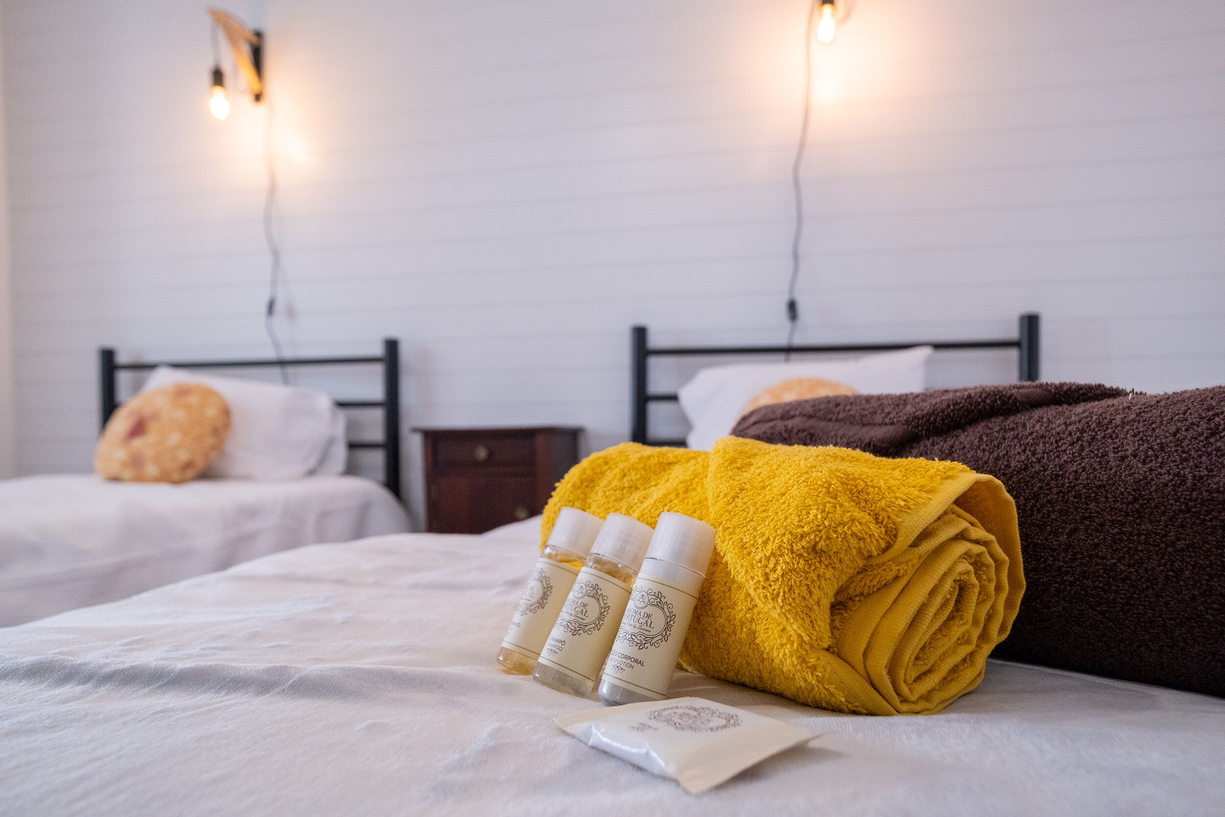 Ein Hotelzimmer mit zwei Betten, jeweils mit Kissen und einem zusammengerollten Handtuch. Auf dem Bett stehen drei Flaschen, und ein kleiner Nachttisch mit einer Lampe.