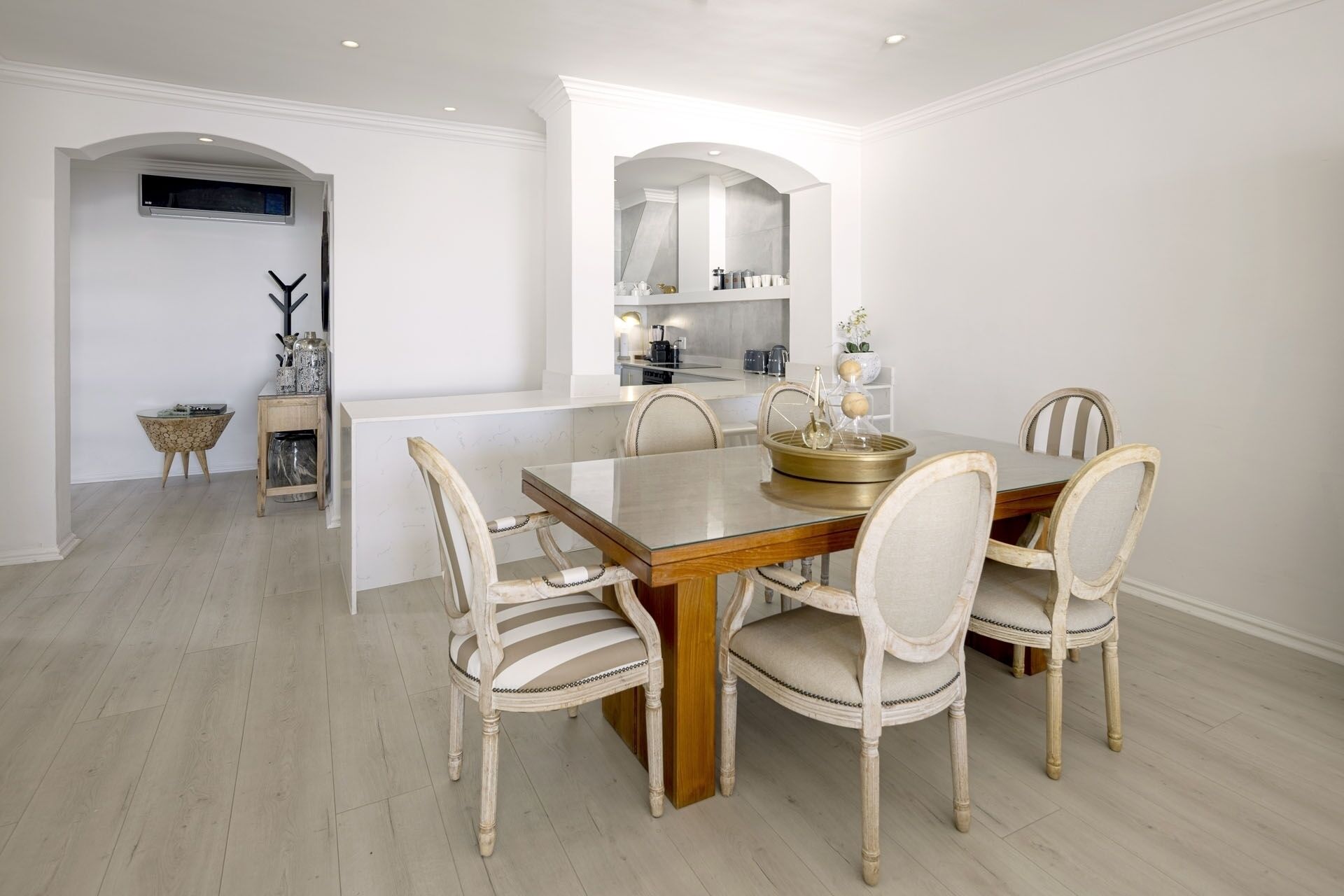 Un comedor con una mesa de madera y seis sillas, una cocina al fondo y una mesita con una planta.