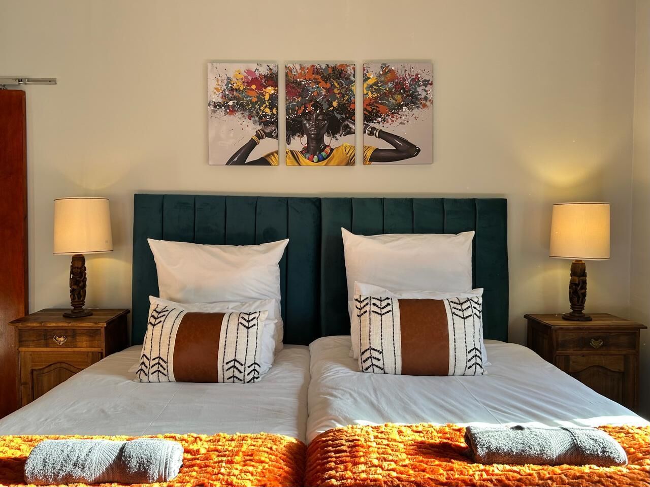 Ein Doppelbett mit weißen Leinentüchern, zwei Kissen mit gemusterten Designs und einer farbenfrohen Decke. Zwei Nachttische mit Lampen und ein an der Wand befestigtes Kunstwerk befinden sich über dem Bett.